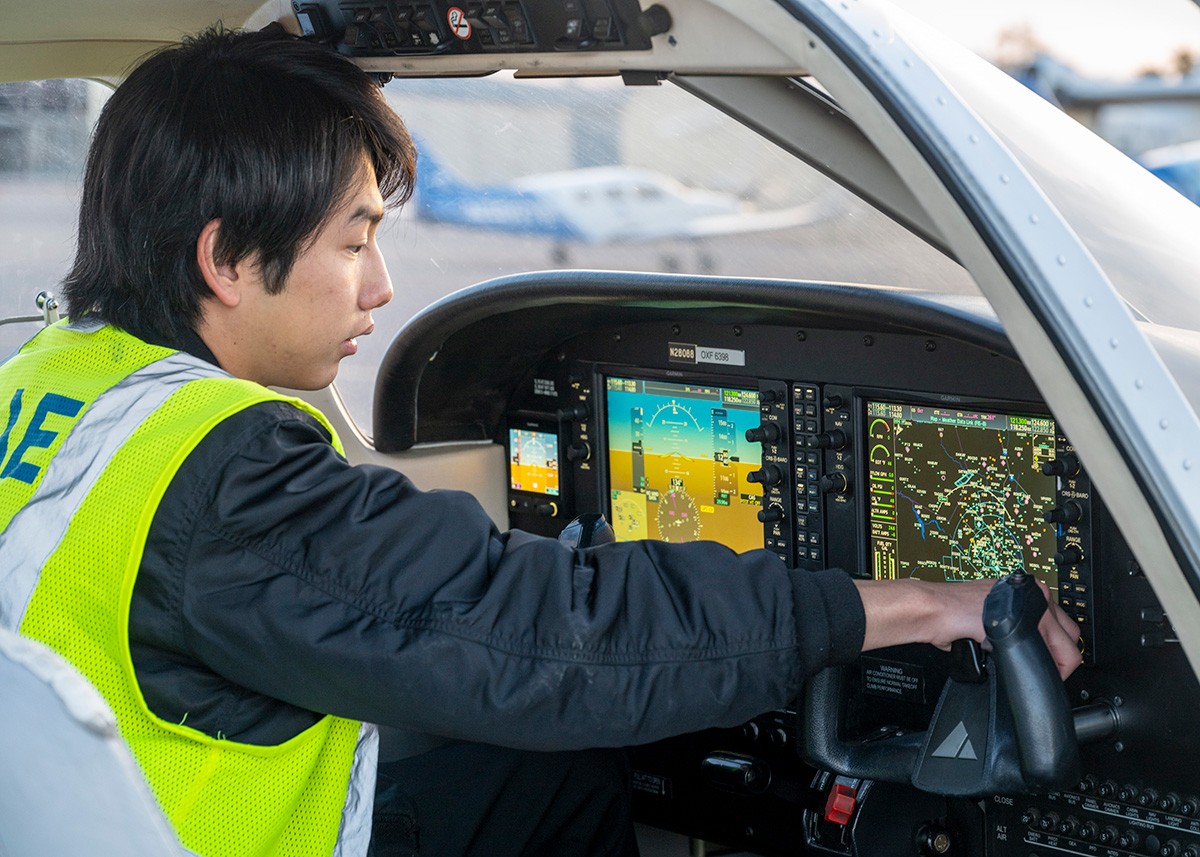 グラスコックピット（Garmin G1000搭載機）の前で計器を操作しながら飛行訓練を行うCAEベスト着用のパイロット訓練生の画像