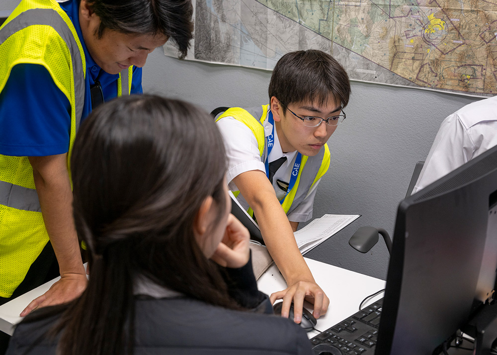 航空地図が貼られた教室で、CAEの教官が訓練生にコンピューター画面を見せながら個別指導を行う様子の画像